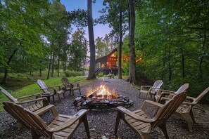 A fire pit surrounded by Adirondack chairs in a forested area with a lodge in the background.