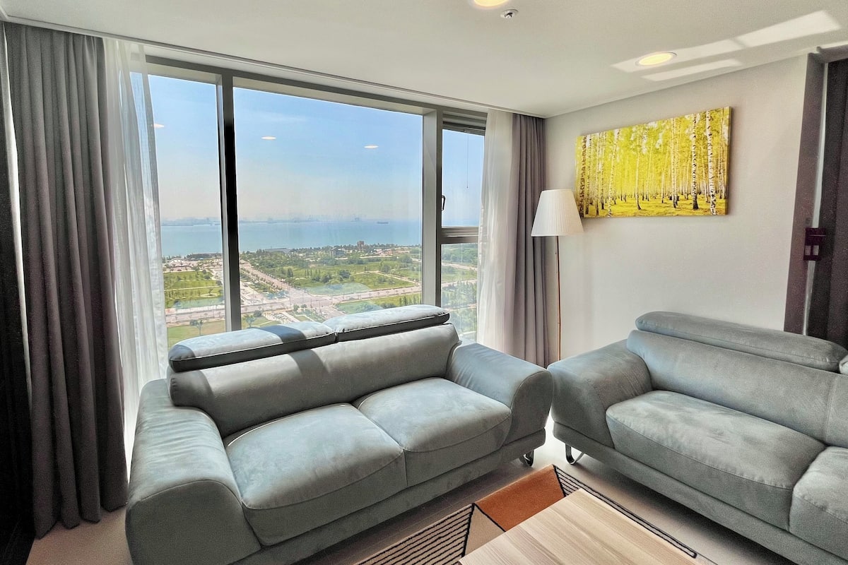 Ein modernes Hotelzimmer mit zwei grauen Sofas, einem großen Fenster mit Blick auf die Stadt und einem Bild an der Wand.