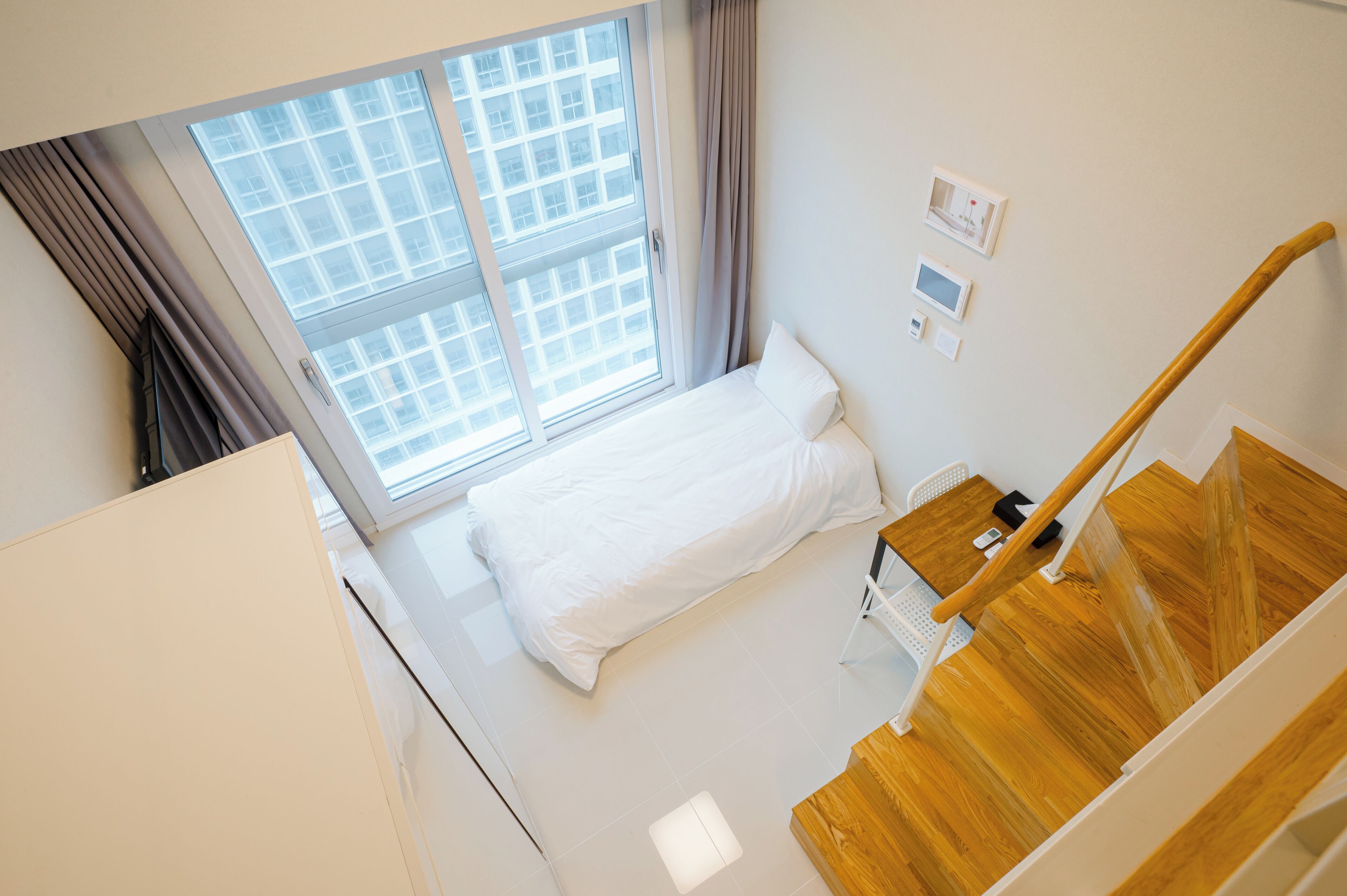 A modern hotel room with a large window, a bed, a wooden staircase, and a small table.