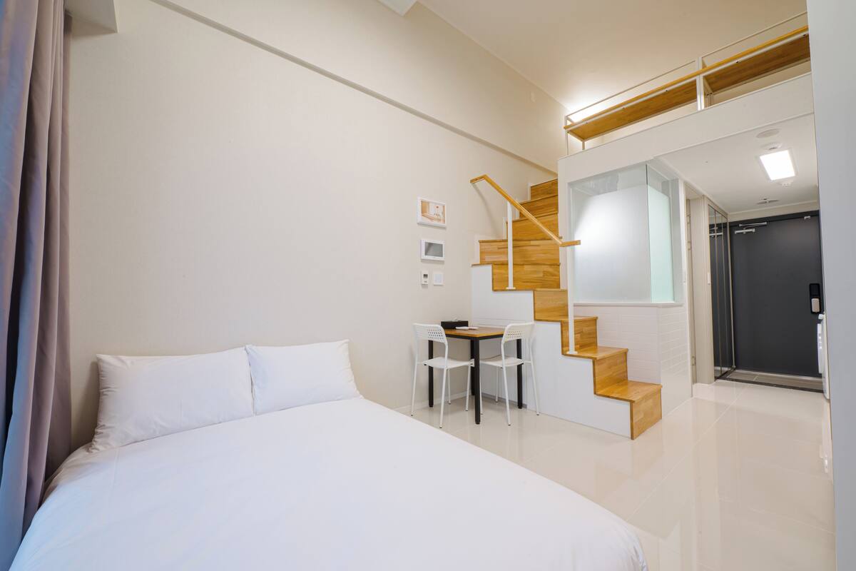 Ein modernes Hotelzimmer mit einem großen Bett, einem kleinen Schreibtisch mit zwei Stühlen, einer Holztreppe und einer Tür zu einem Badezimmer.