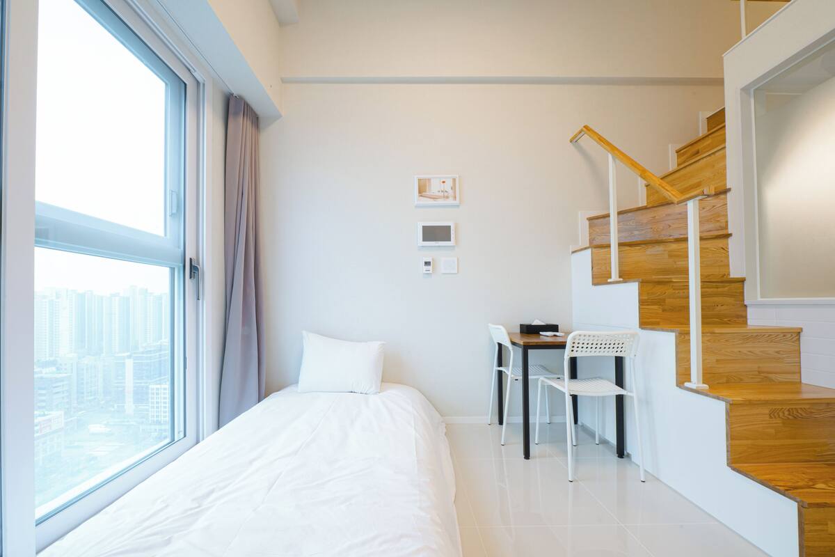 Ein modernes Hotelzimmer mit einem großen Bett, einem Schreibtisch mit zwei Stühlen, einer Holztreppe und Blick auf die Stadt durch das Fenster.