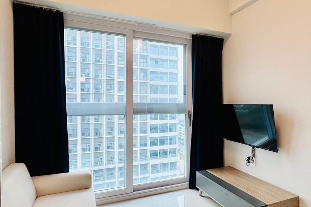 Ein modernes Wohnzimmer mit einer Couch, einem Fernseher auf einem Schrank und einem großen Fenster mit Vorhängen.