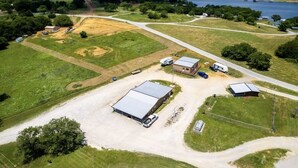 Vue aérienne d’une propriété rurale comprenant plusieurs bâtiments, dont une grande grange et des constructions plus petites, le tout entouré de champs ouverts et bordé d’une route.