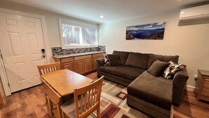 Ein gemütlicher Wohnbereich mit Küchenzeile, einem Essbereich mit Holztisch und -stühlen, einem braunen Sofa mit Kissen und einem Teppichboden.