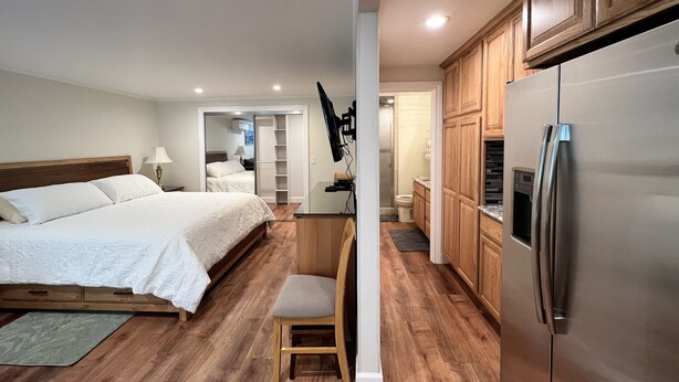 Ein Schlafzimmer mit einem Bett, Nachttisch und einem Sessel. Eine Küchenzeile mit Kühlschrank und Holzschränken ist ebenfalls vorhanden. Durch eine geöffnete Tür ist ein Badezimmer zu sehen.