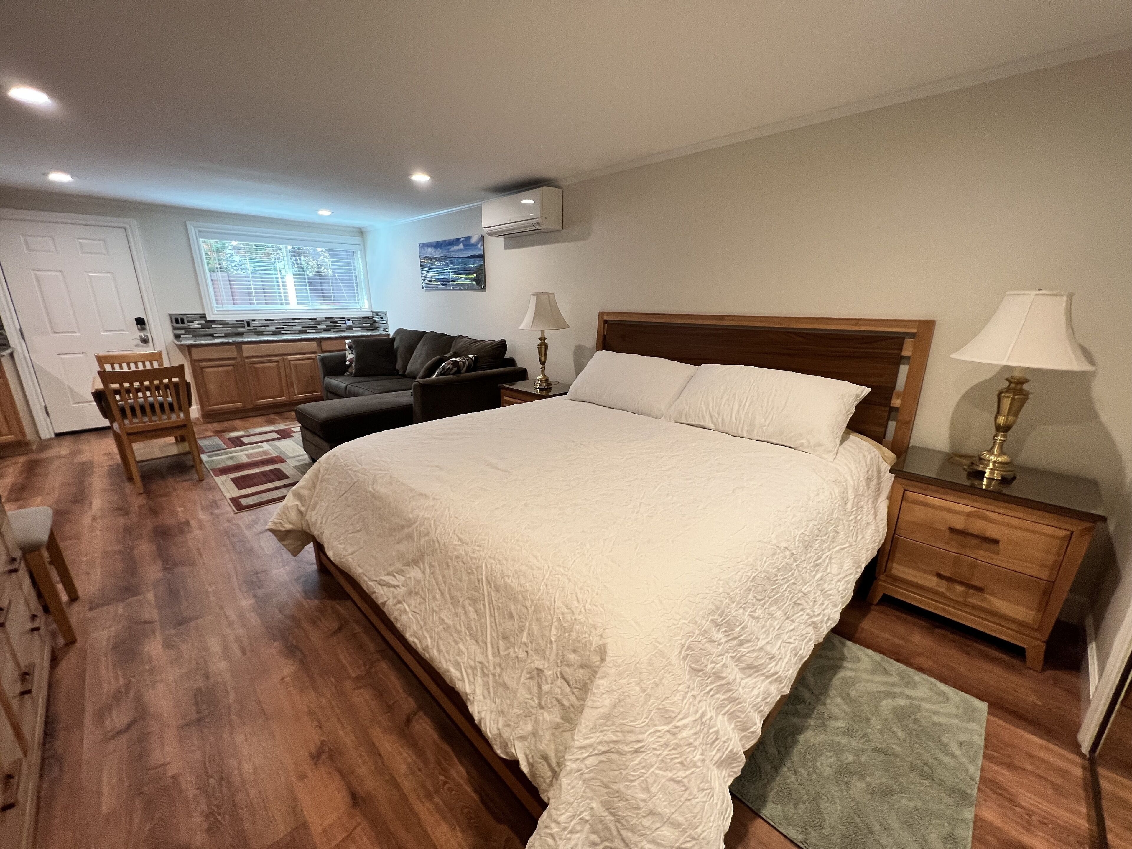 A bedroom with a large bed, wooden headboard, bedside tables with lamps, a nightstand, a chair, a small table, and a window with blinds.