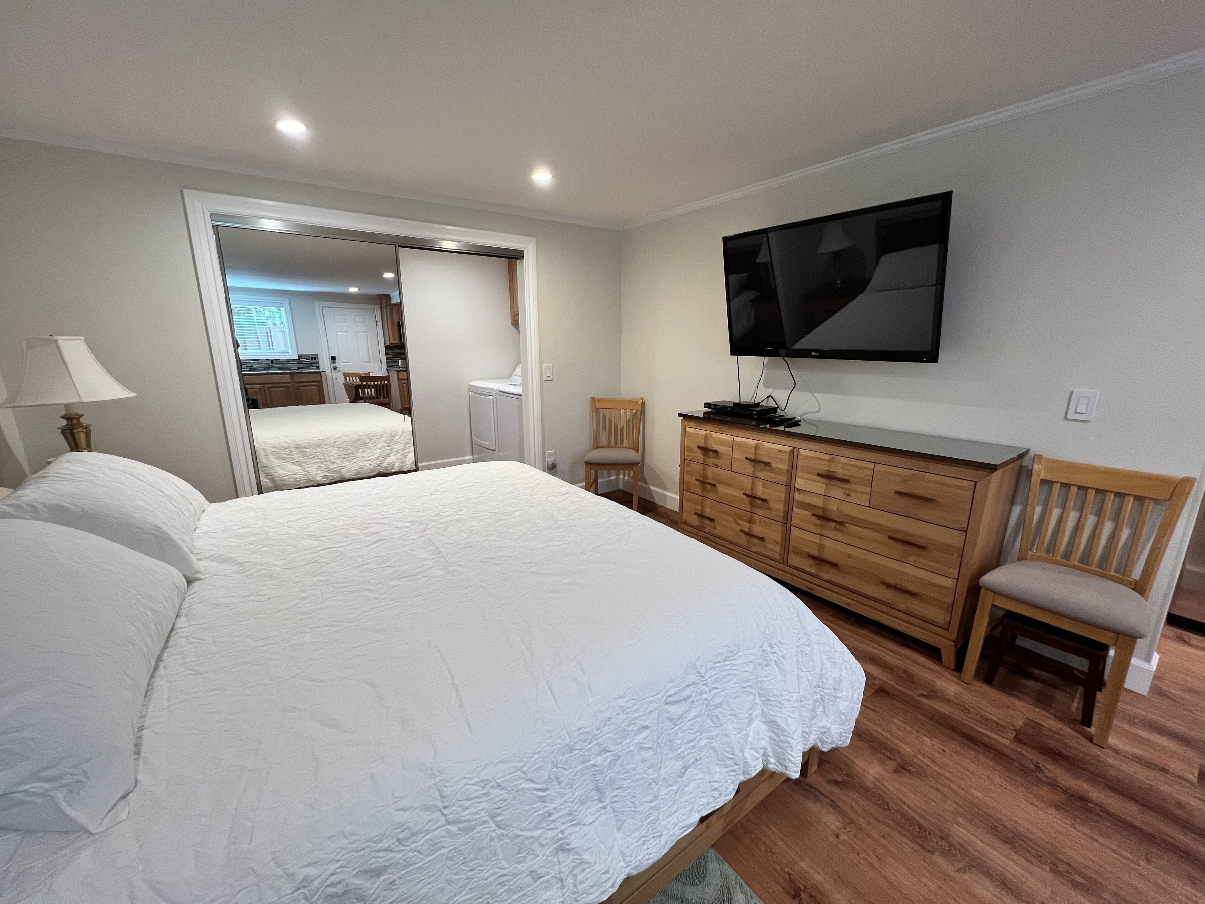 A bedroom with a large bed, a wooden dresser, a TV mounted on the wall, and a mirror reflecting the room.