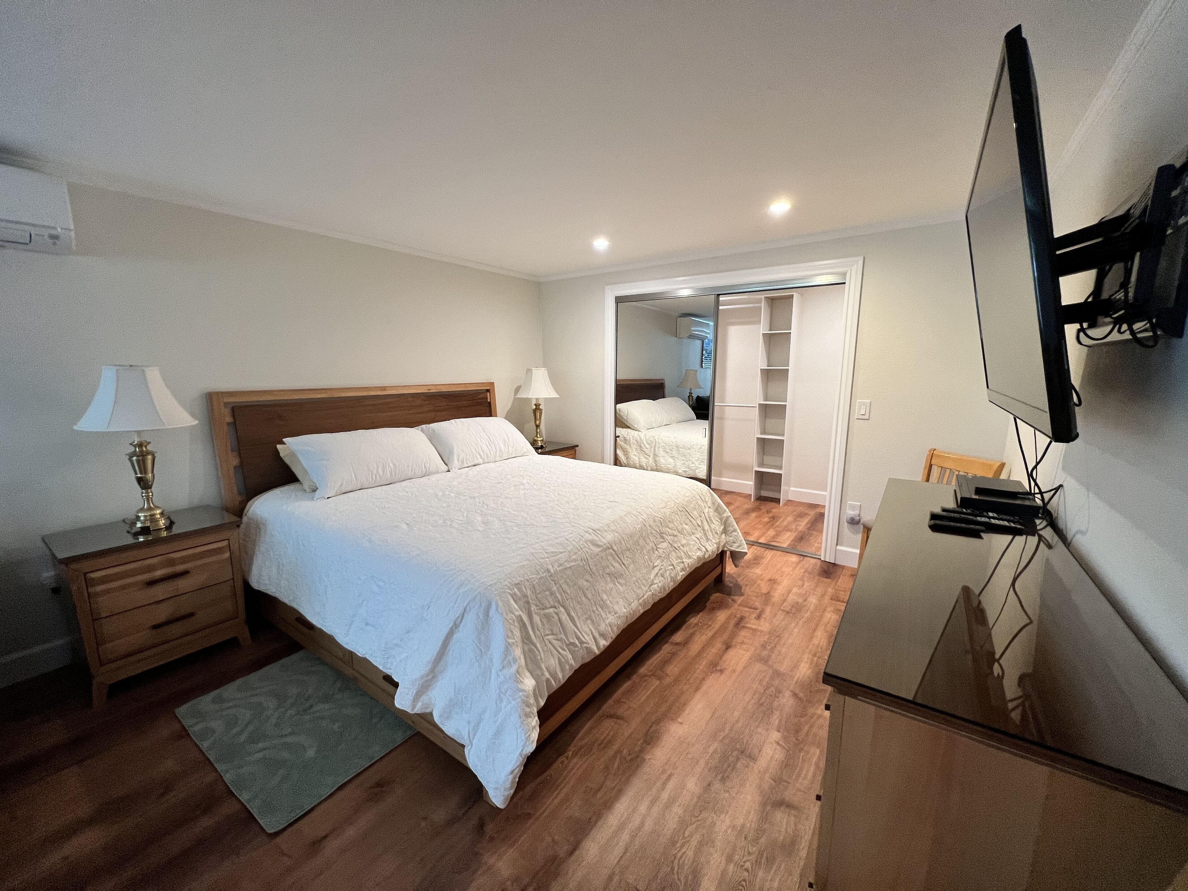 A bedroom with a wooden bed, bedside tables, a mirror, and a TV mounted on the wall.