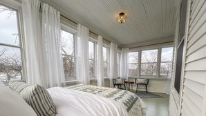 A bedroom with a large bed, a desk, and two chairs. There are multiple windows providing natural light and views of the outside.