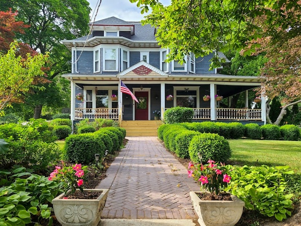 A two-story house with a front porch, a flag, and a well-maintained garden with potted plants and flowers.