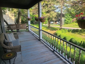 A covered porch with a wooden bench and wicker furniture, overlooking a well-maintained garden and a road.