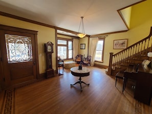 A spacious living room with hardwood floors, a large wooden staircase, and a grand chandelier.