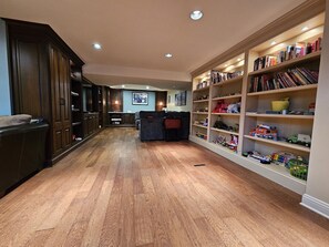 A spacious living room with a wooden floor, a dark wood cabinet, a grey sofa, and a bookshelf filled with books and toys.