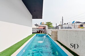 A swimming pool with a water feature, surrounded by a white wall and a small patch of green grass.