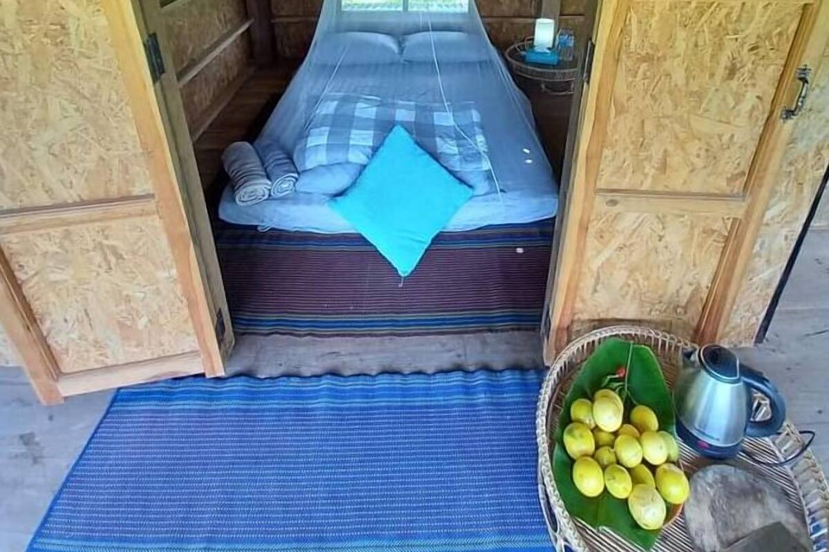 A single bed with a mosquito net, a blue mat, and a basket of fruit on the floor.