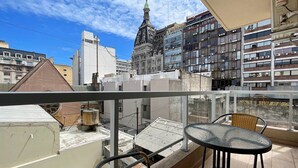 A balcony with wicker chairs and a table, overlooking a cityscape with historic buildings and modern high-rises.