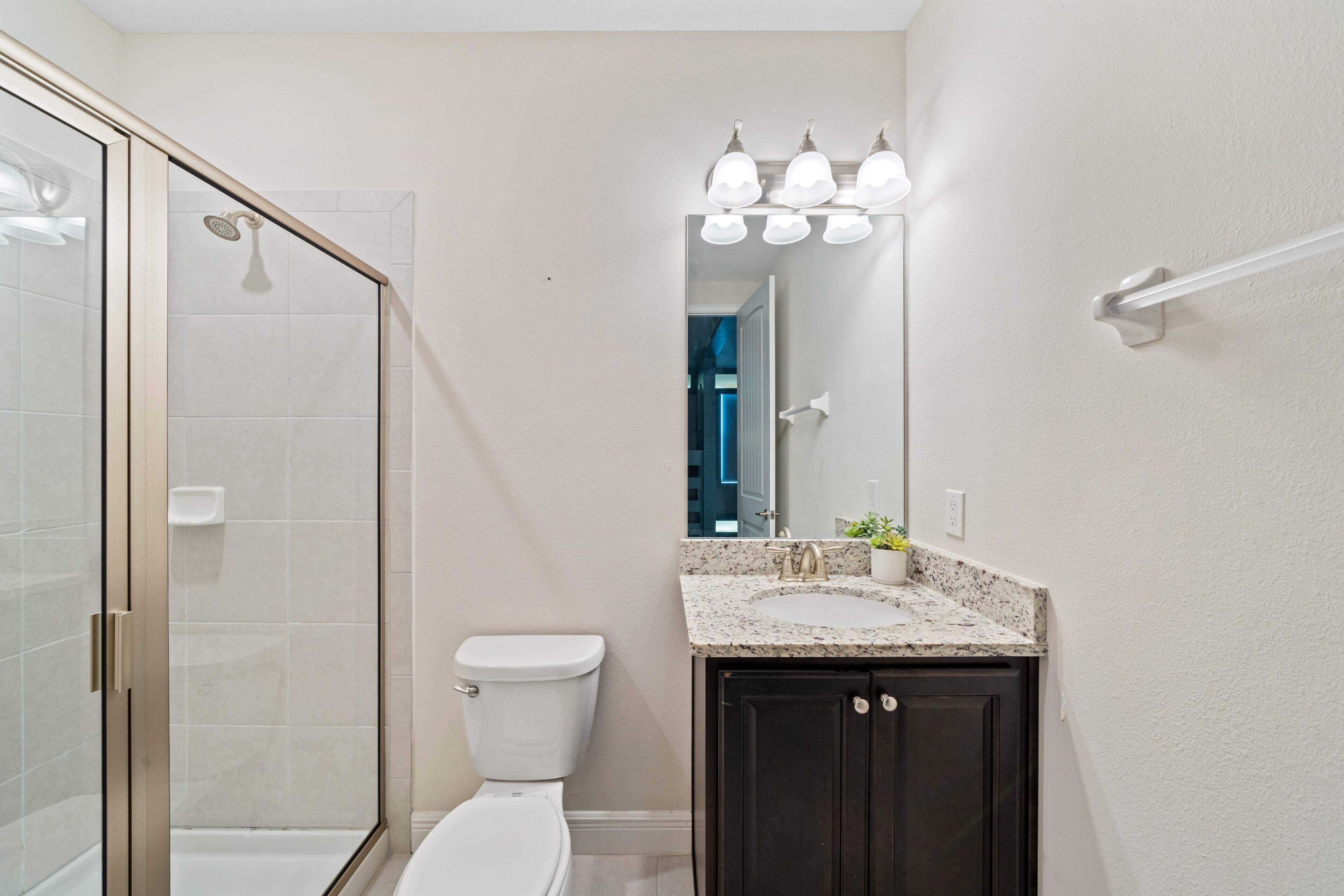 A bathroom with a glass shower enclosure, a white toilet, a dark vanity with a sink, and a mirror with lights above.