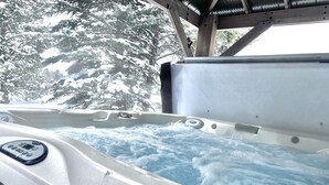 Un bain à remous doté d’un panneau de commande, entouré d’une terrasse en bois et d’arbres couverts de neige.