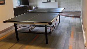 Mesa de pingue-pongue em um quarto com parede de tijolos, televisão e quadro emoldurado.