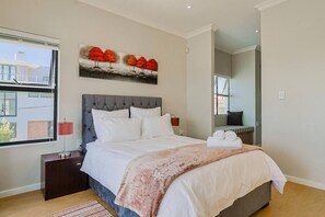 A modern hotel room with a large bed, a painting of trees, a window with a view, and a small bedside table with a lamp.