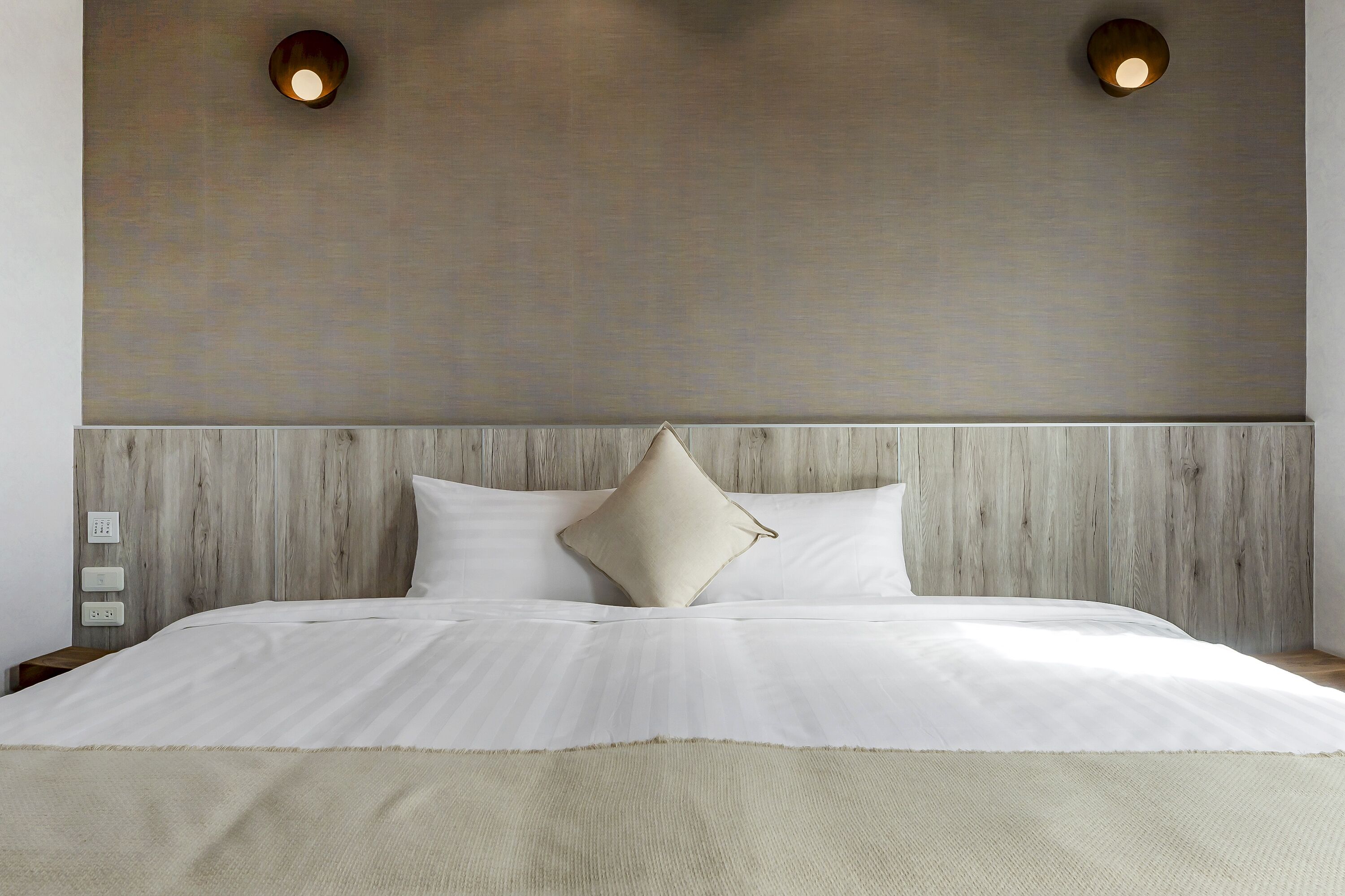 Una cama bien hecha con sábanas blancas y una manta color beige, cabecera de madera y dos lámparas fijadas a la pared.