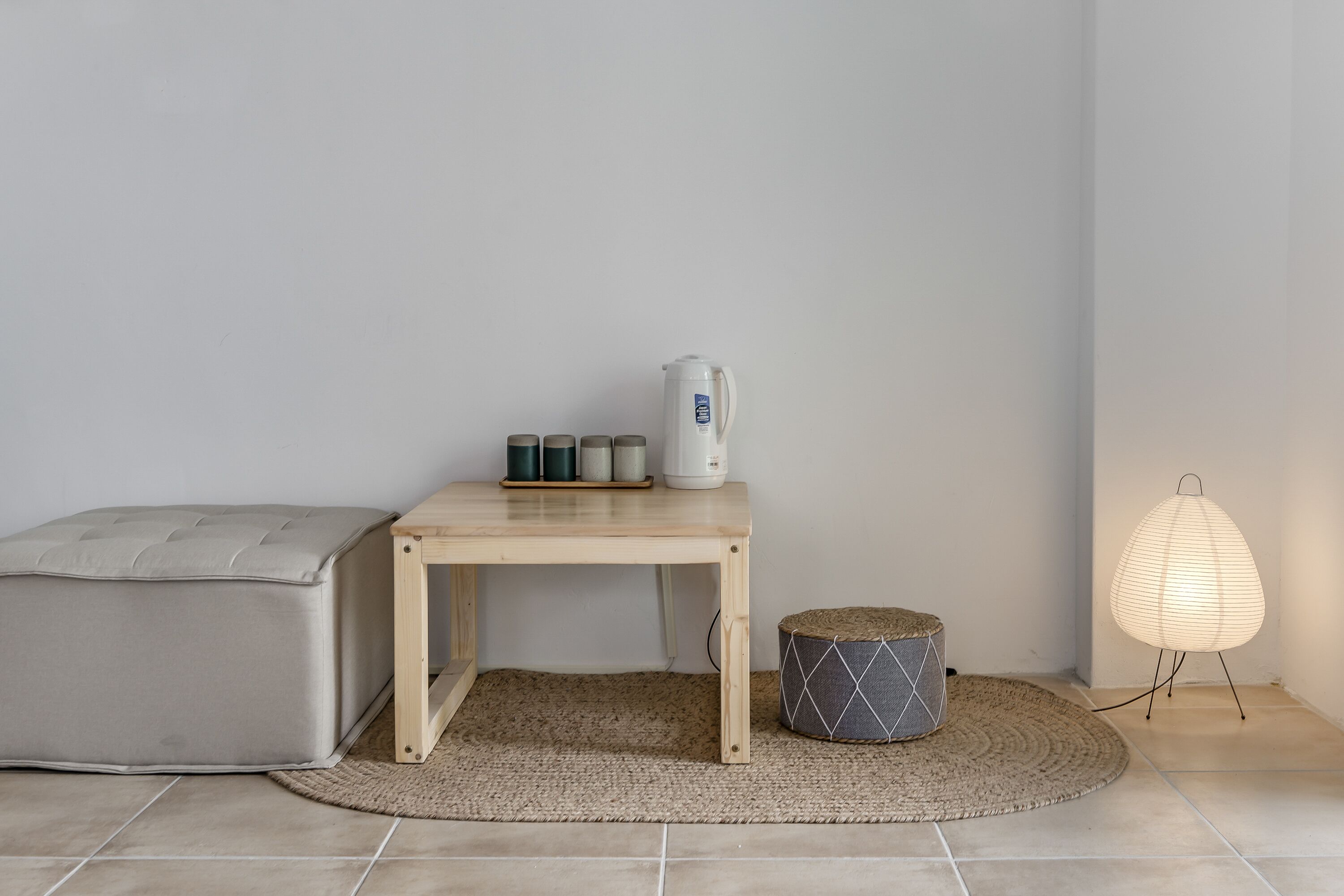 Una habitación minimalista con una mesa de madera, un sofá gris, una alfombra pequeña, una lámpara y un hervidor.