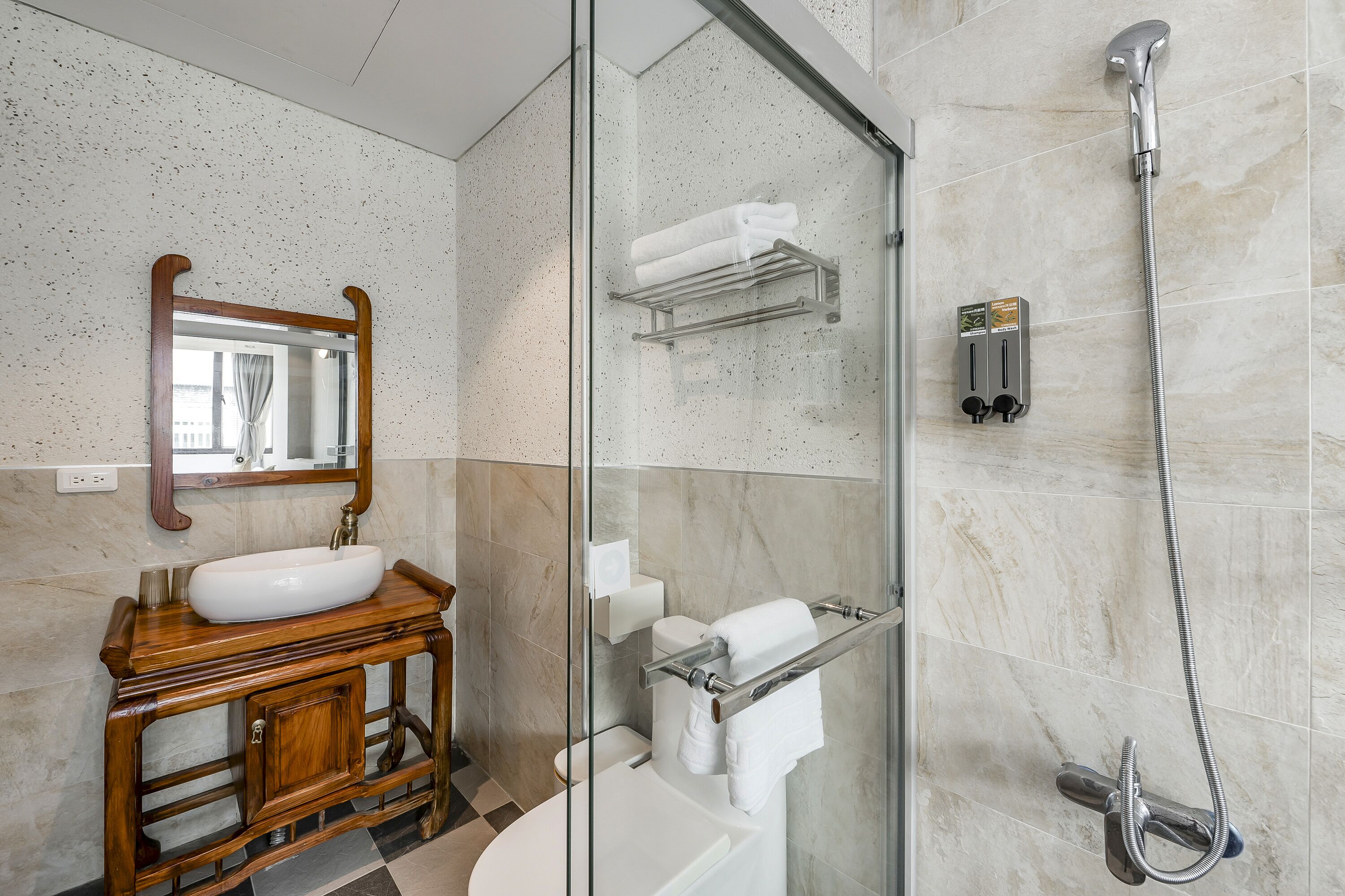 Un baño con tocador de madera, cabina de ducha de vidrio y pared con azulejos de mármol.