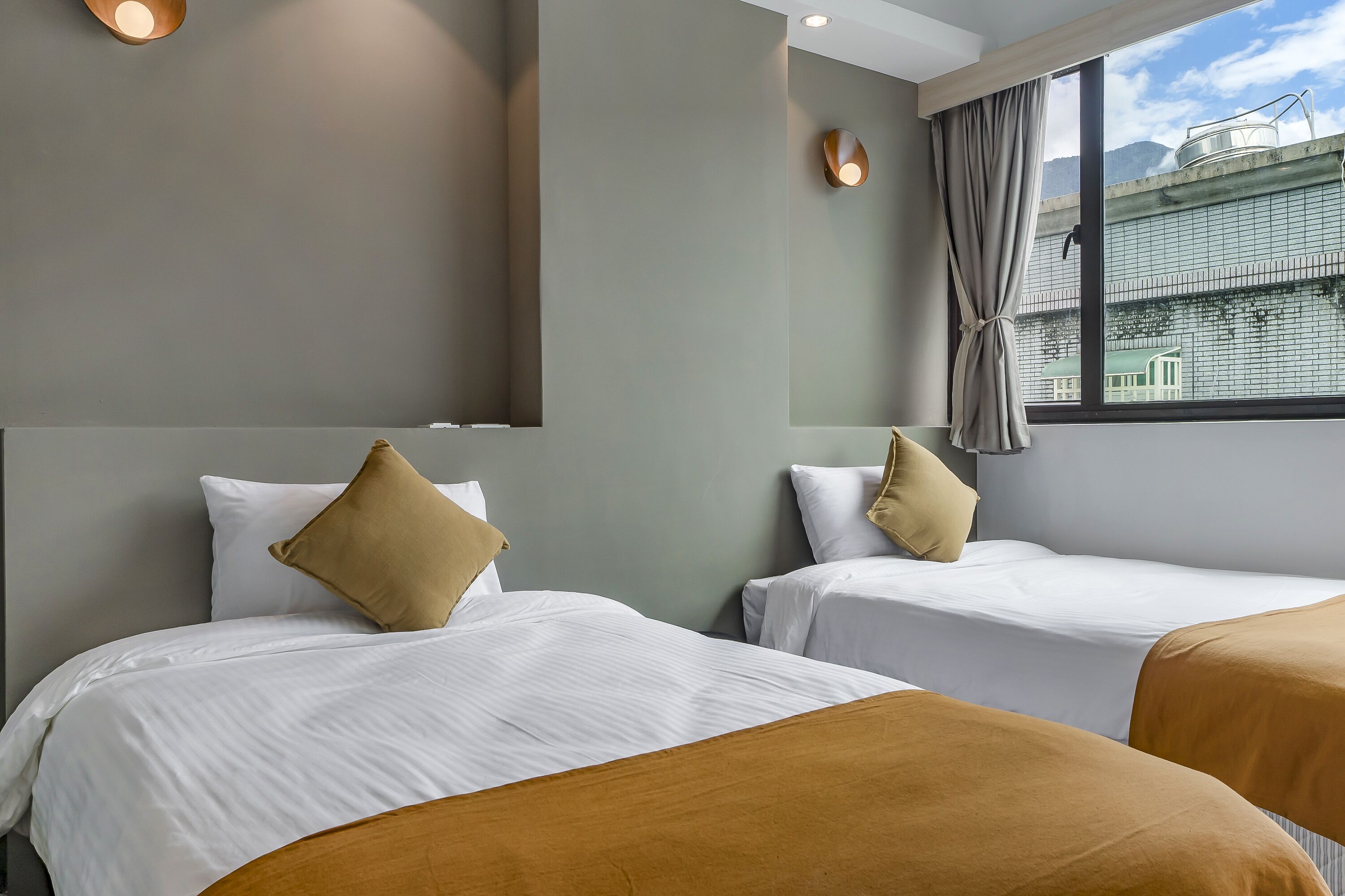 A hotel room with two beds, each with white bedding and a brown accent pillow. The room has a large window with a view of buildings and mountains.