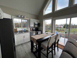 A modern kitchen with white cabinets, a black refrigerator, a dining table with chairs, and large windows offering a view of the outdoors. - Elk Lodge in Sooke (Sooke)