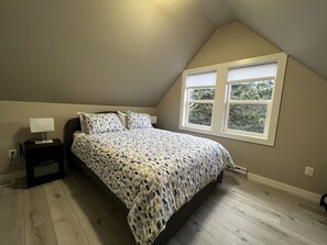 A bedroom with a bed, a nightstand, a lamp, and a window with blinds. - Elk Lodge in Sooke (Sooke)