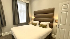 Een net opgemaakt bed met witte lakens en twee kussens, een groot gestoffeerd hoofdbord en een bedlampje.