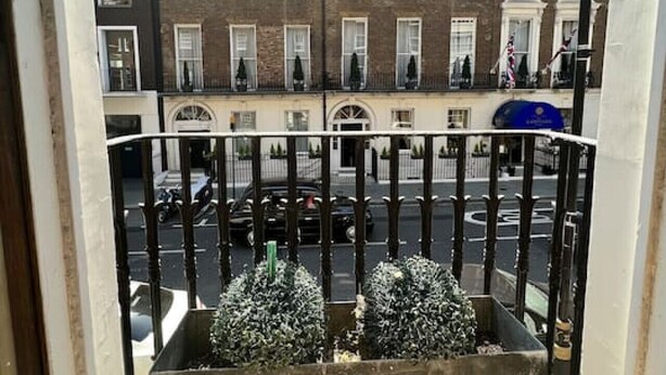Een balkon met een metalen hekwerk, uitzicht op een straat met geparkeerde auto's en een rij bakstenen gebouwen met meerdere ramen en balkons.