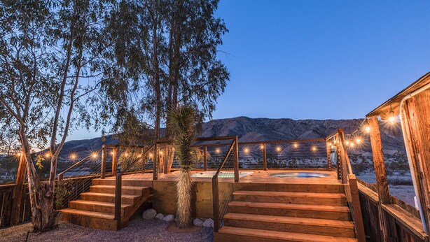 A wooden deck with a hot tub, surrounded by a wooden fence and string lights. - Desert Playground - hot tub, dipping pool, hammocks, stargazing, dog friendly! (Twentynine Palms)