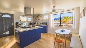 A compact kitchen with a central island, a dining area with a small table and chairs, and a large window offering a view of a desert landscape. - Desert Playground - hot tub, dipping pool, hammocks, stargazing, dog friendly! (Twentynine Palms)