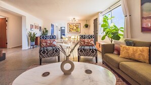 A modern living room with a round coffee table, two patterned armchairs, a sofa with cushions, a dining area with a table and chairs, and a large window with curtains. - Desert Playground - hot tub, dipping pool, hammocks, stargazing, dog friendly! (Twentynine Palms)