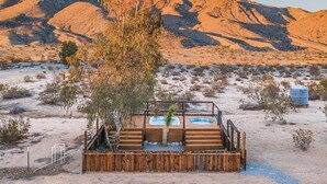 A wooden deck with a hot tub and a small pool, surrounded by a wooden fence, set against a desert landscape with sparse vegetation and mountains in the background. - Desert Playground - hot tub, dipping pool, hammocks, stargazing, dog friendly! (Twentynine Palms)