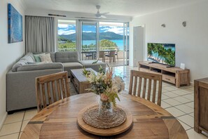 A modern living room with a sofa, a dining table with chairs, a TV, and a view of the ocean.