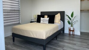 Un dormitorio moderno con un cabecero oscuro, una cama con cubrecama color beige, una almohada con el texto 'welcome home', una mesita de noche con una lámpara y una planta en maceta.