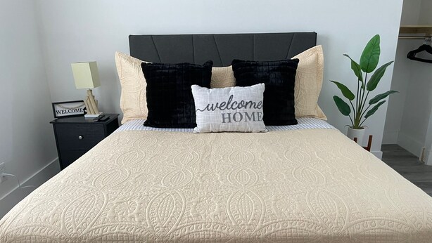 Una cama bien hecha con colcha color crema, almohadas negras y color crema, y una almohada con la inscripción 'bienvenido a casa'.