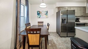 A dining area with a wooden table and chairs, a stainless steel refrigerator, and a window with blinds.