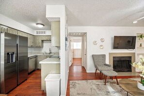 A modern kitchen with stainless steel appliances, a dining area with a grey upholstered chair, and a living room with a fireplace and a flat-screen TV. - I51 - Apt|Patio|11mi to Convention Ctr|Sleeps 2 (Irving)
