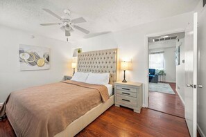 A bedroom with a tufted headboard, a bed with a brown blanket, bedside lamps, a ceiling fan, and a door leading to another room.