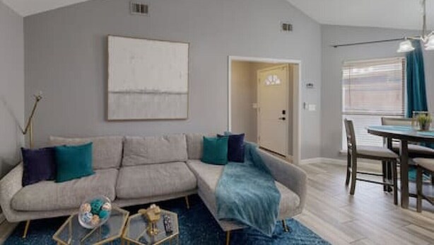 A modern living room with a grey sofa, teal cushions, a coffee table, and a dining area with a table and chairs. - Modern Comfort Lux Stay | Near DFW Airport (Euless)