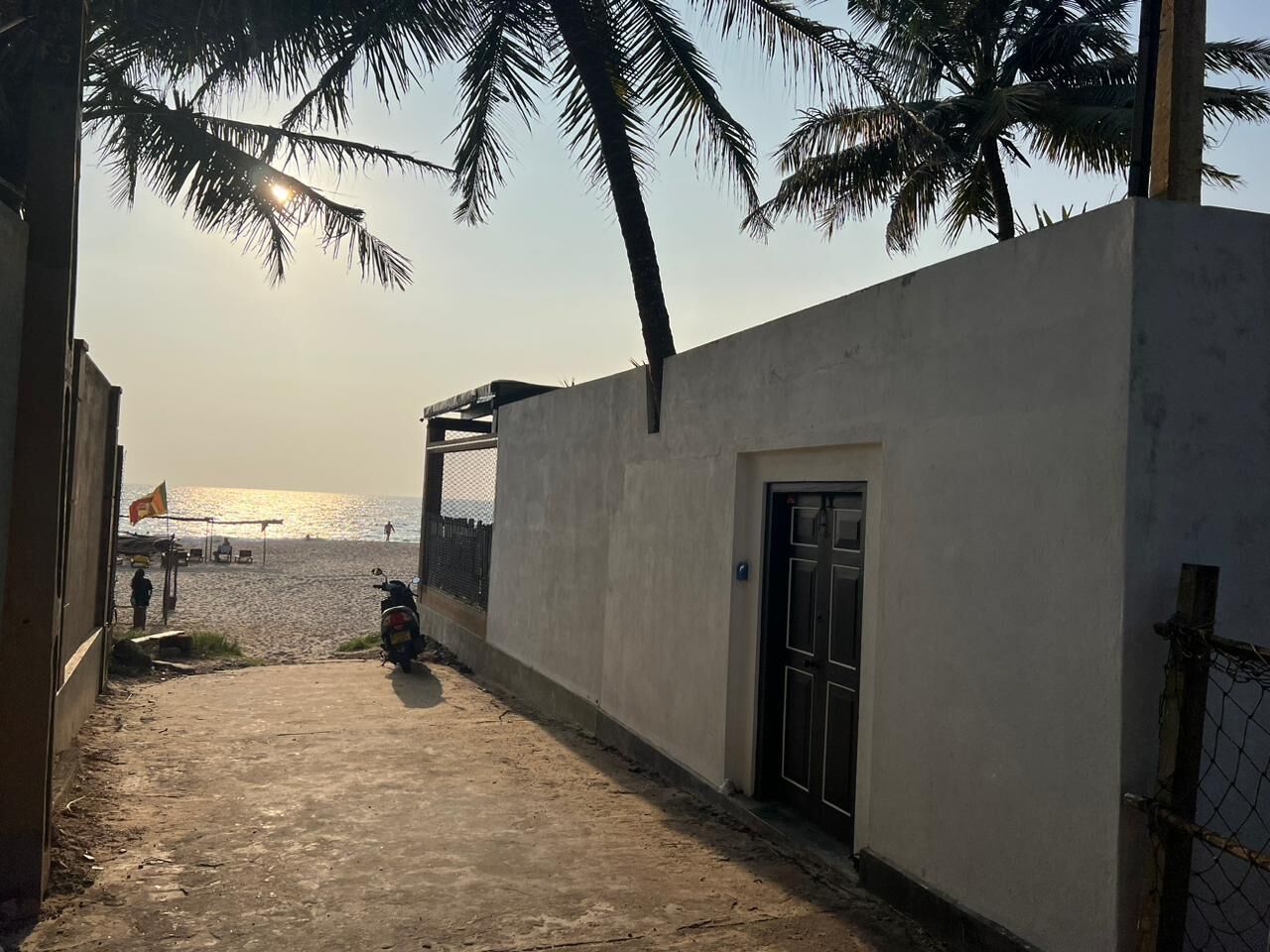 A narrow concrete pathway leading to a building with a door, palm trees, and a view of the sea.