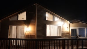 Une maison en bois à deux étages, dotée d’un balcon, de grandes fenêtres et d’un éclairage extérieur.