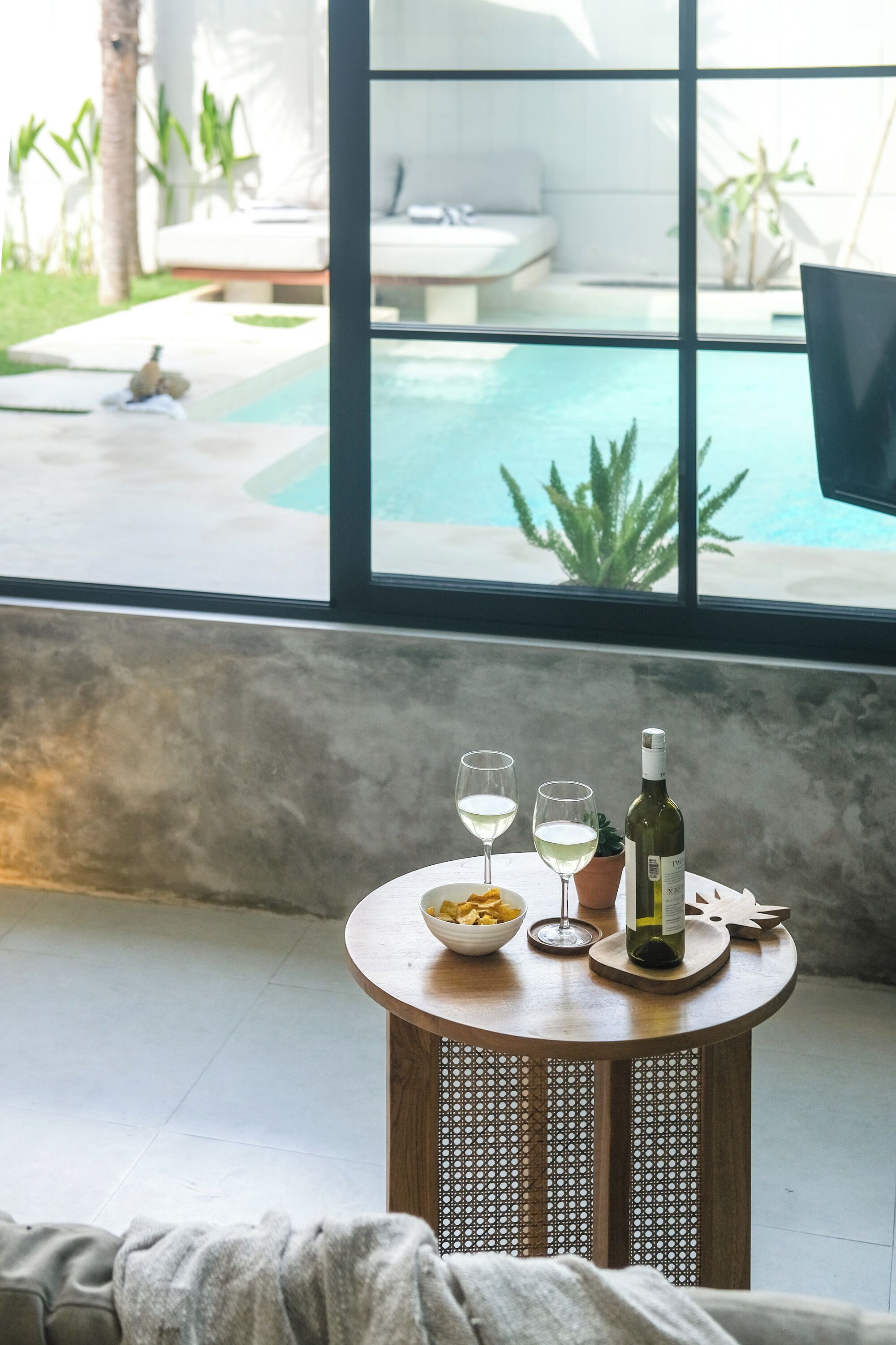 A round wooden table with a bowl of fruit, two wine glasses, and a bottle of wine. A large window reveals a pool and outdoor seating area.