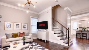A modern living room with a sofa, coffee table, and a flat-screen TV displaying 'NETFLIX'. A staircase leads to a kitchen area.