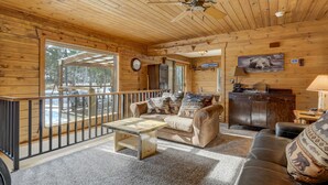 A cozy cabin interior with a wooden ceiling, a sofa with animal-themed cushions, a coffee table, a dining area with a wooden cabinet, and a large window offering a view of the outdoors. - The Bear Den  (Florissant)