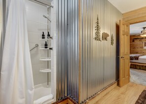A bathroom with a shower, a toilet, and a sink with shelves. There is a wooden door leading to a bedroom. - The Bear Den  (Florissant)