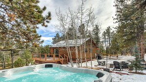 A hot tub surrounded by snow-covered ground, wooden deck, and a cabin with a deck and outdoor seating. - The Bear Den  (Florissant)